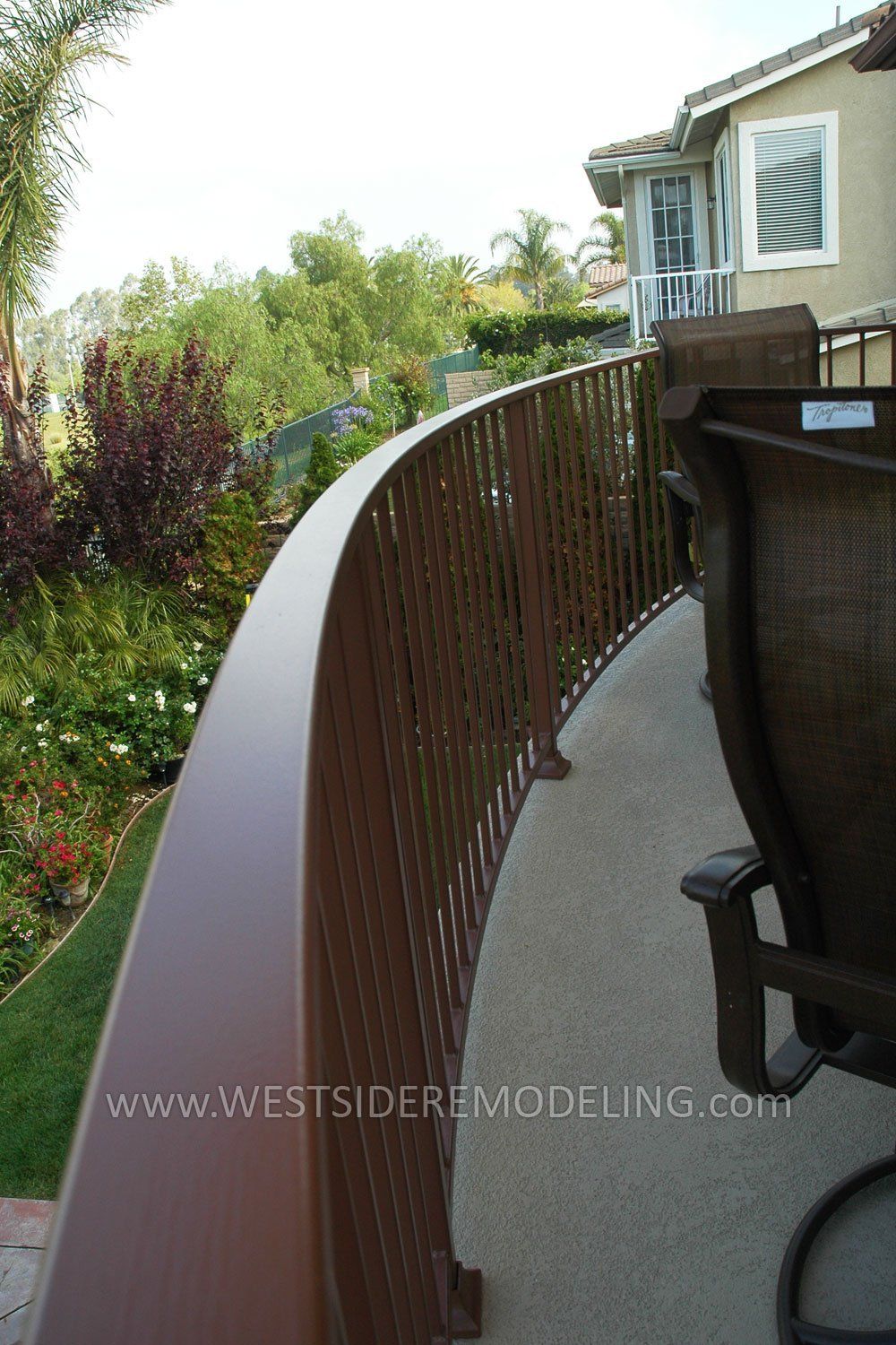 Curved brown deck railing along a narrow patio beside a house and garden path