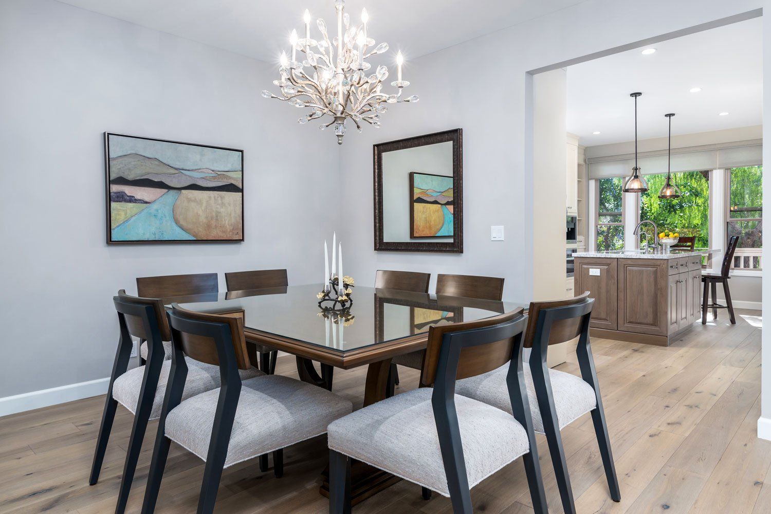 Bright dining room with a glass table, dark chairs, chandelier, and open view to a kitchen and garden.