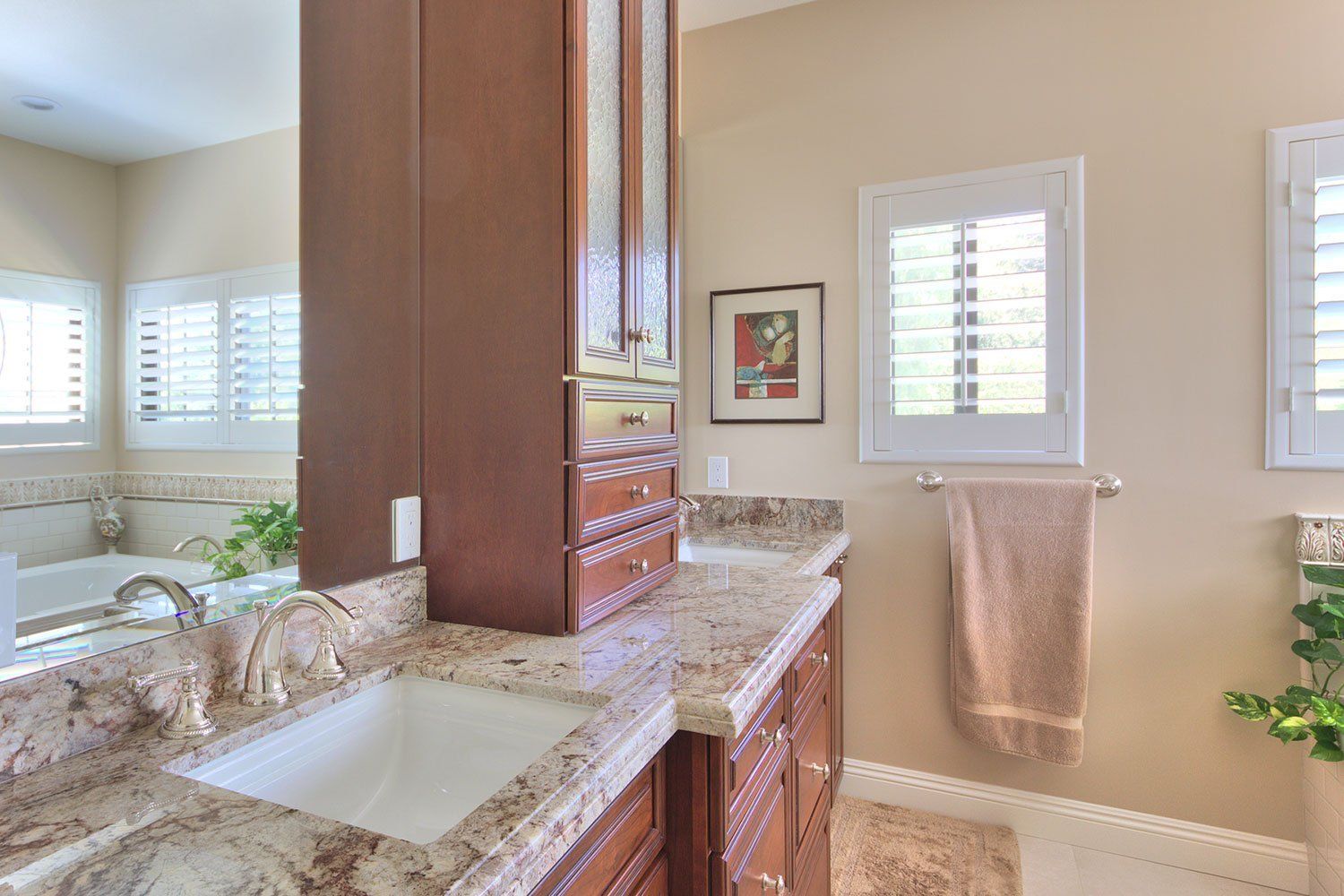 Bright bathroom with granite double vanity, wood cabinets, wall mirror, and window with towel rack
