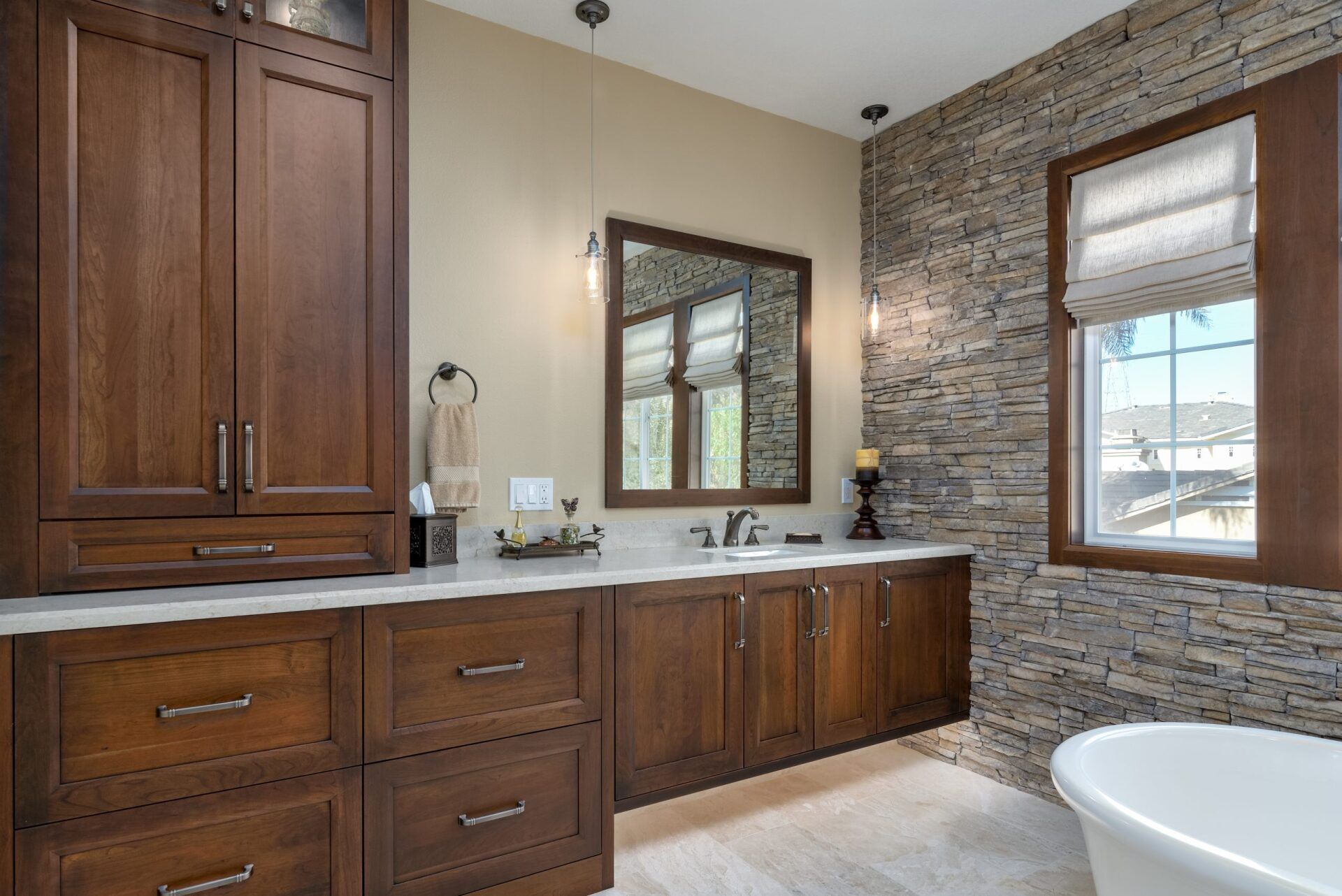 Modern bathroom vanity with dark wood cabinets, stone accent wall, mirror, and bathtub by a window.