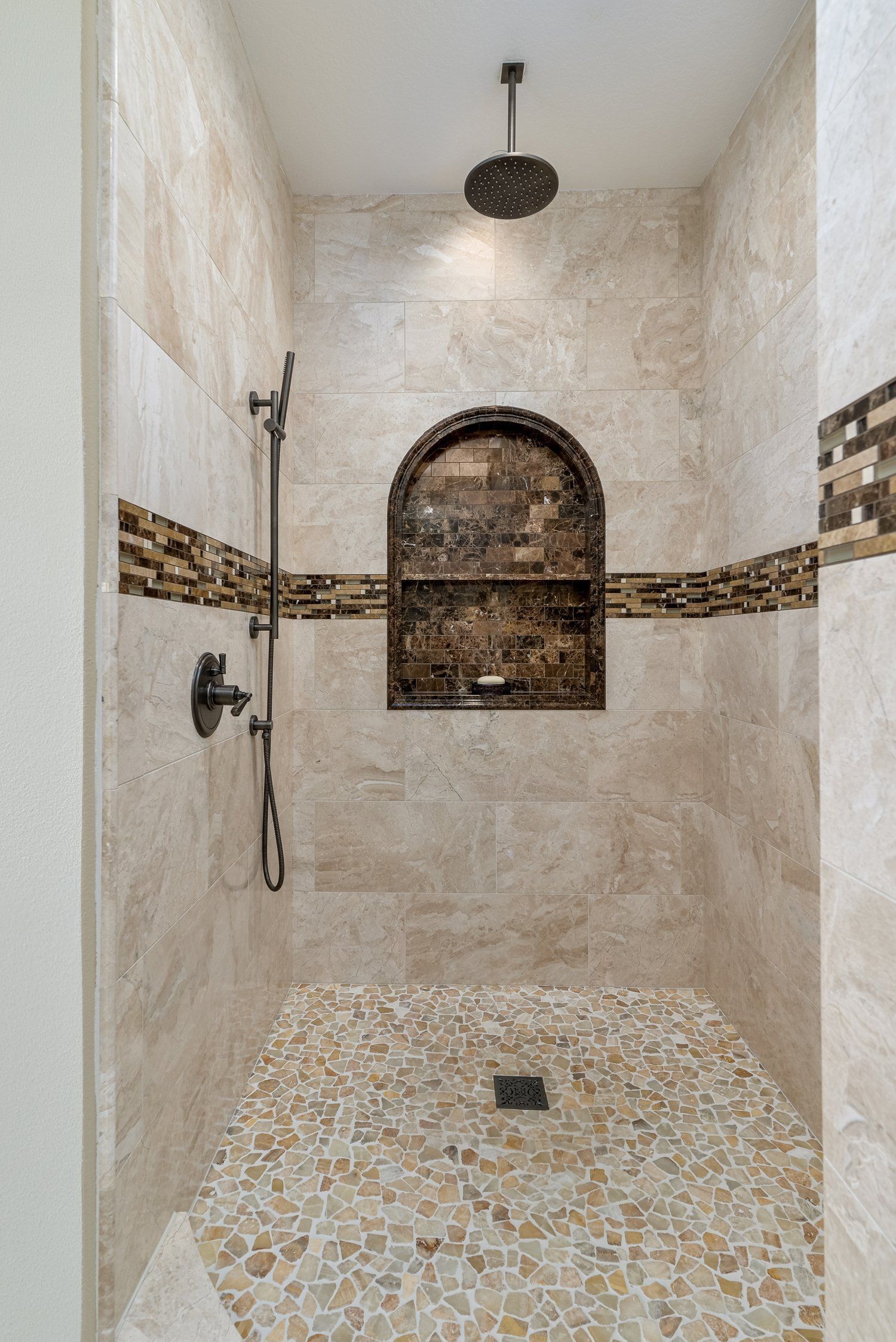 Empty tiled shower with arched niche, handheld showerhead, and mosaic floor.