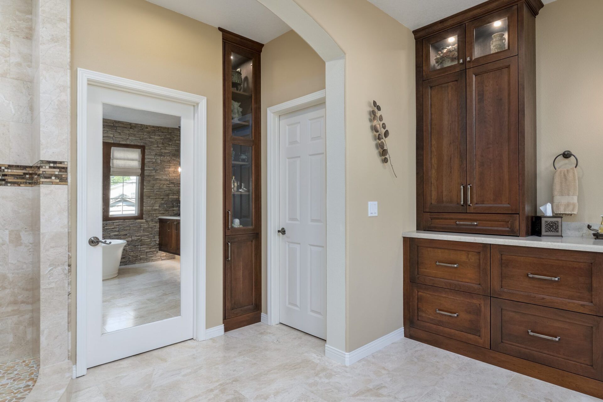 Bright bathroom vanity area with wood cabinets, white tile floor, and a large mirror reflecting a bedroom.