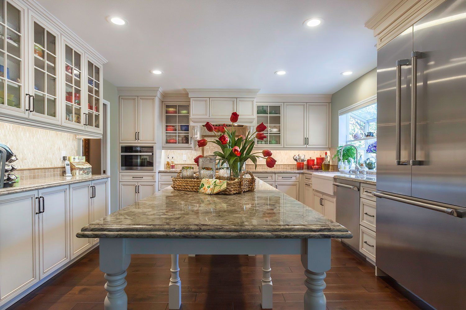Bright modern kitchen with white cabinets, stainless steel fridge, and a large granite island with flowers.