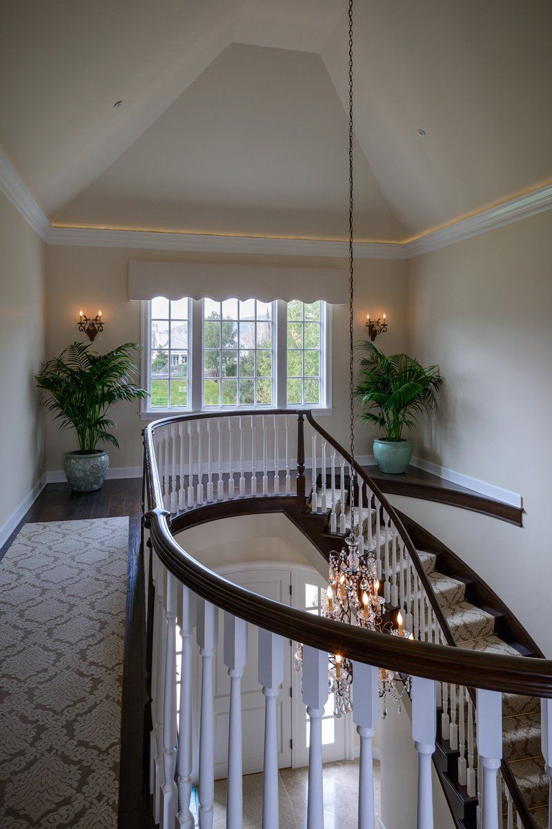 Elegant stair landing with curved railing, chandelier, potted plants, and a large window in a bright hallway