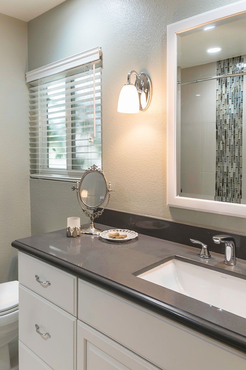 Modern bathroom vanity with dark countertop, white cabinets, mirror, and window blinds