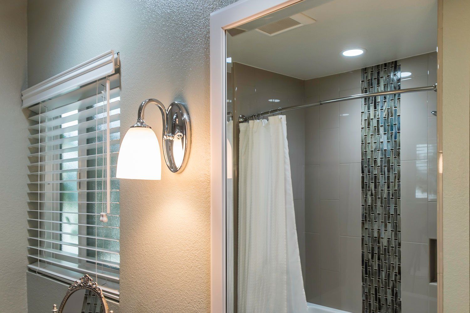 Bathroom vanity mirror beside a lit wall sconce, window blinds, and shower curtain in a bright tiled bathroom