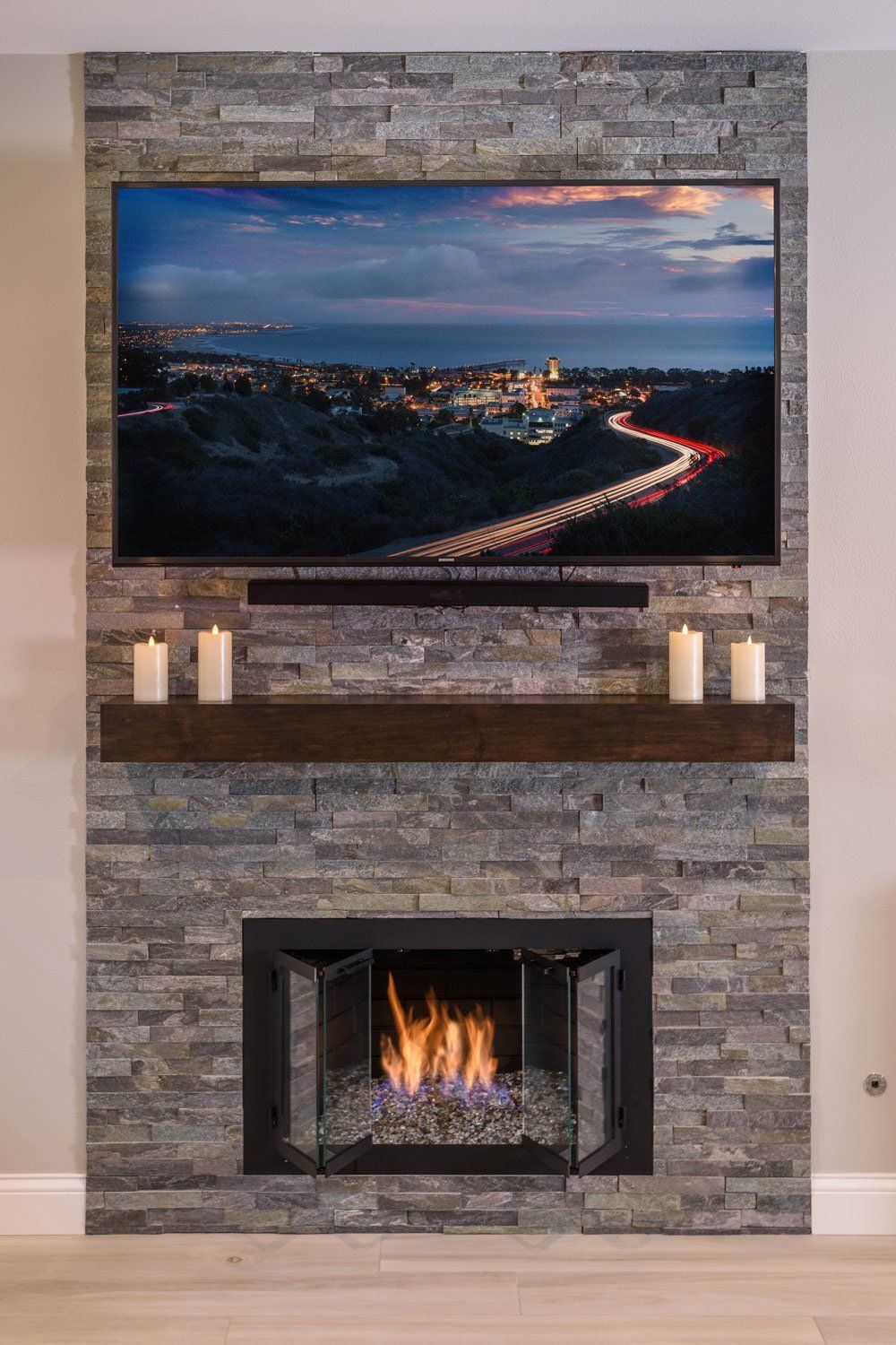 Living room stone fireplace with a lit fire and a wall-mounted TV above it showing a cityscape at dusk