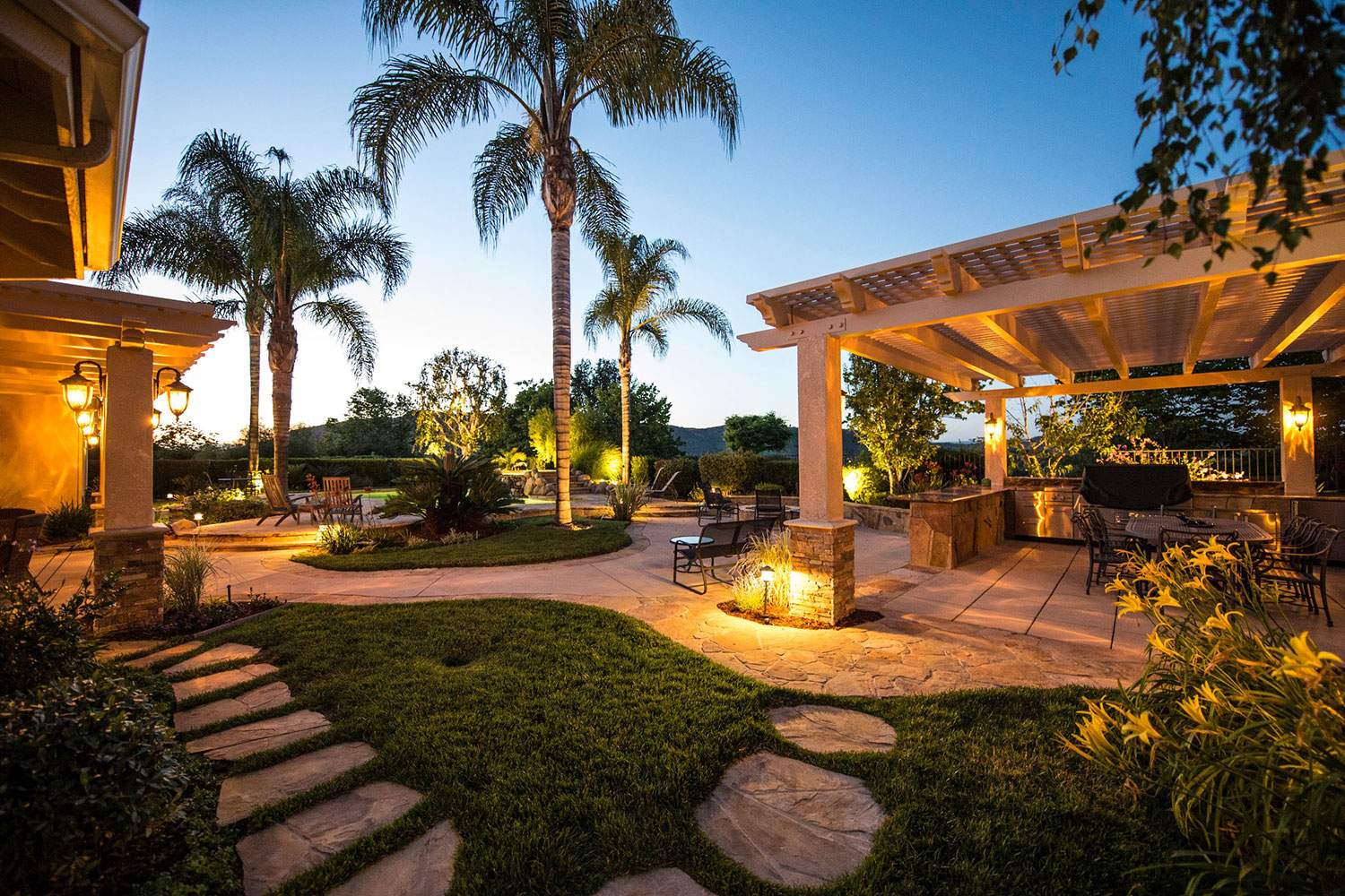 Tropical backyard patio at sunset with palm trees, stone path, warm lights, and a covered seating area