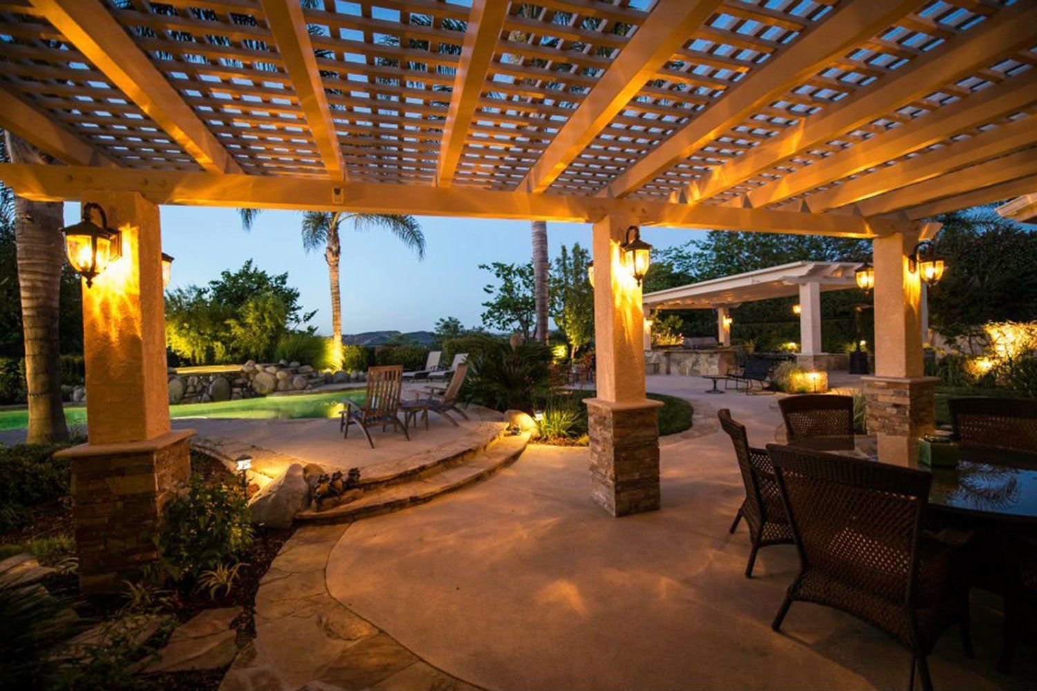 Covered patio at dusk with wicker seating, warm lights, and a garden view beyond the open pergola.