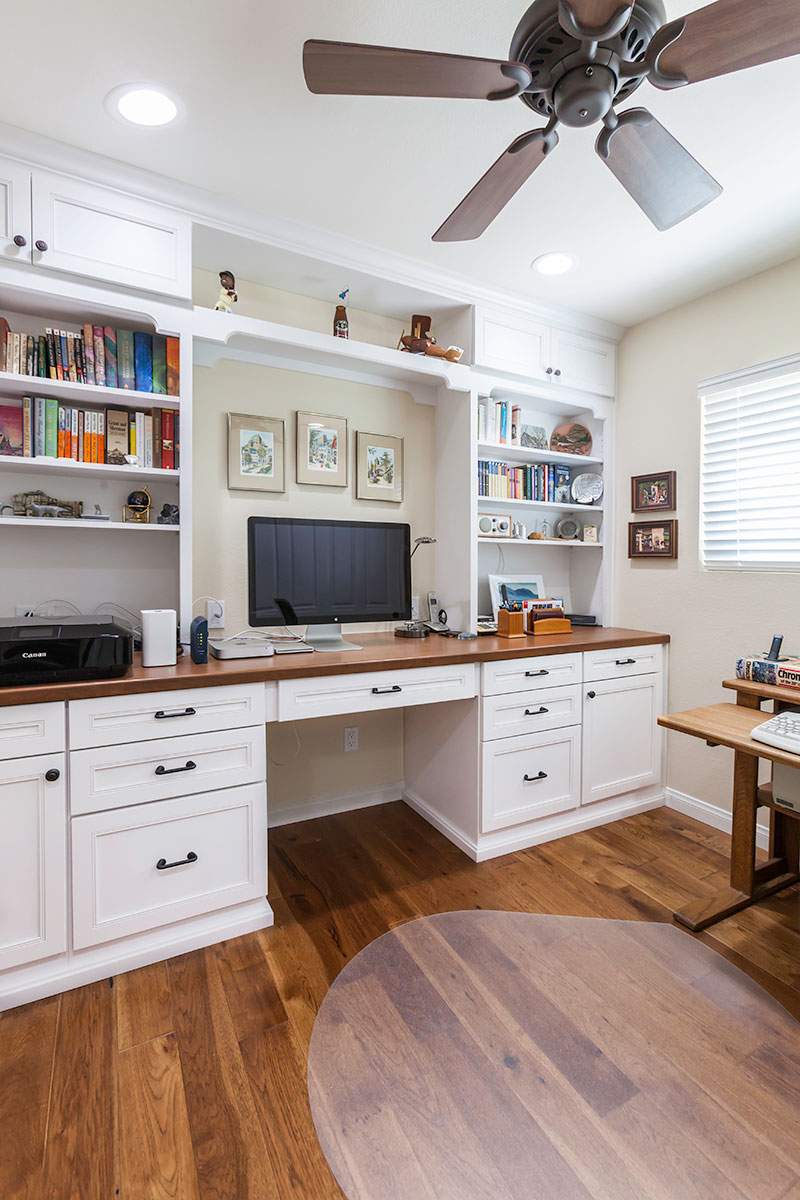 Bright home office with white built-in desk, TV, bookshelves, wooden floor, and ceiling fan