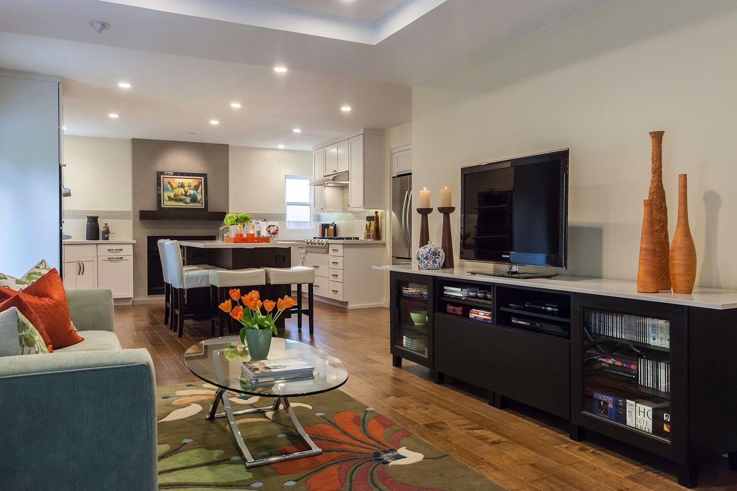 Modern open-plan living room and kitchen with TV, black console, glass coffee table, and warm wood floors.