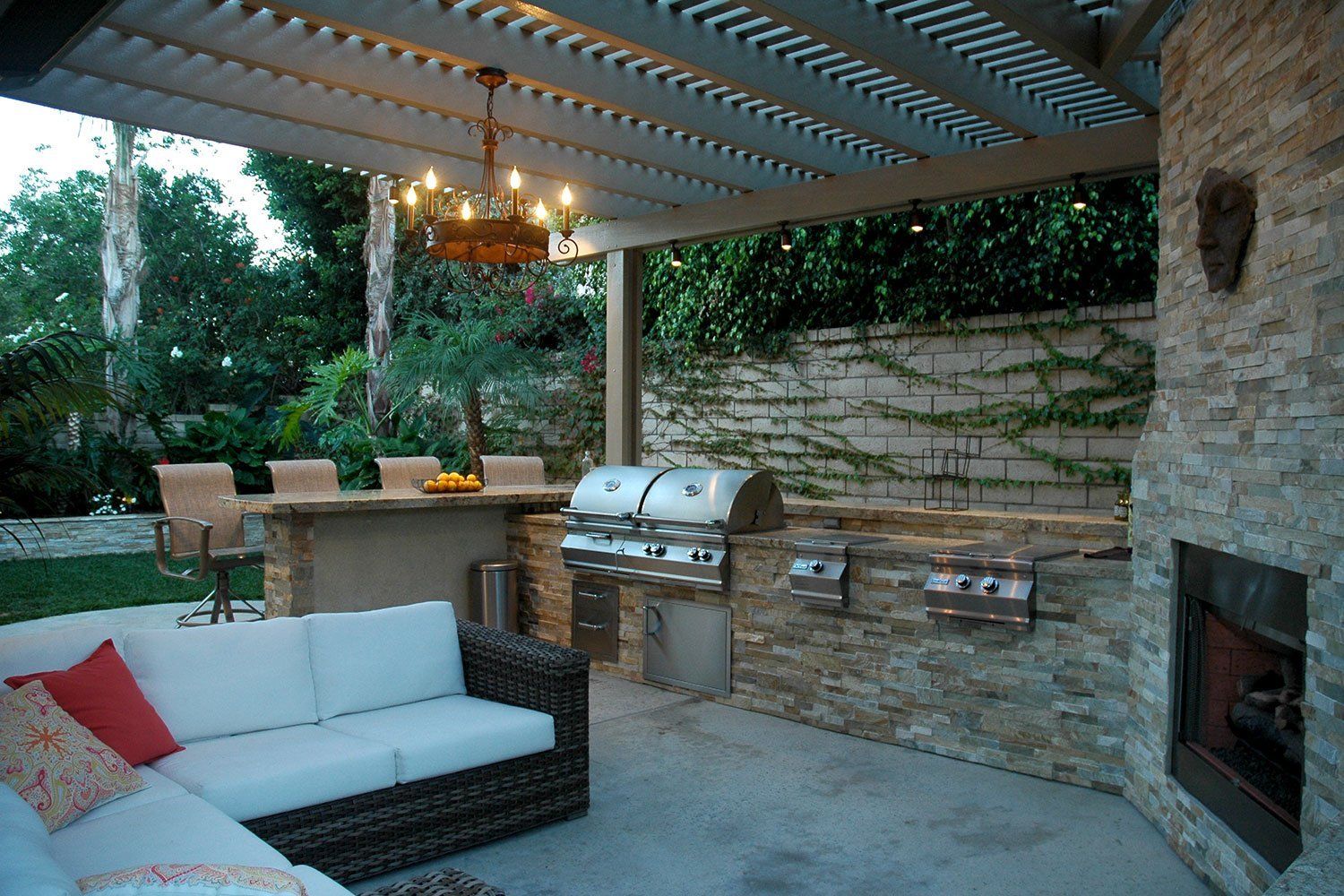Outdoor patio with white sofa, stone fireplace, grill, and dining table under a slatted pergola