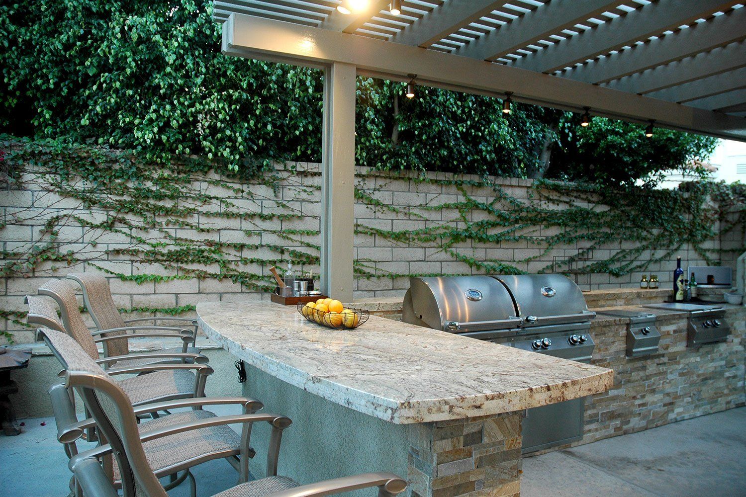 Covered outdoor patio with stone table, chairs, and built-in grill under a slatted pergola