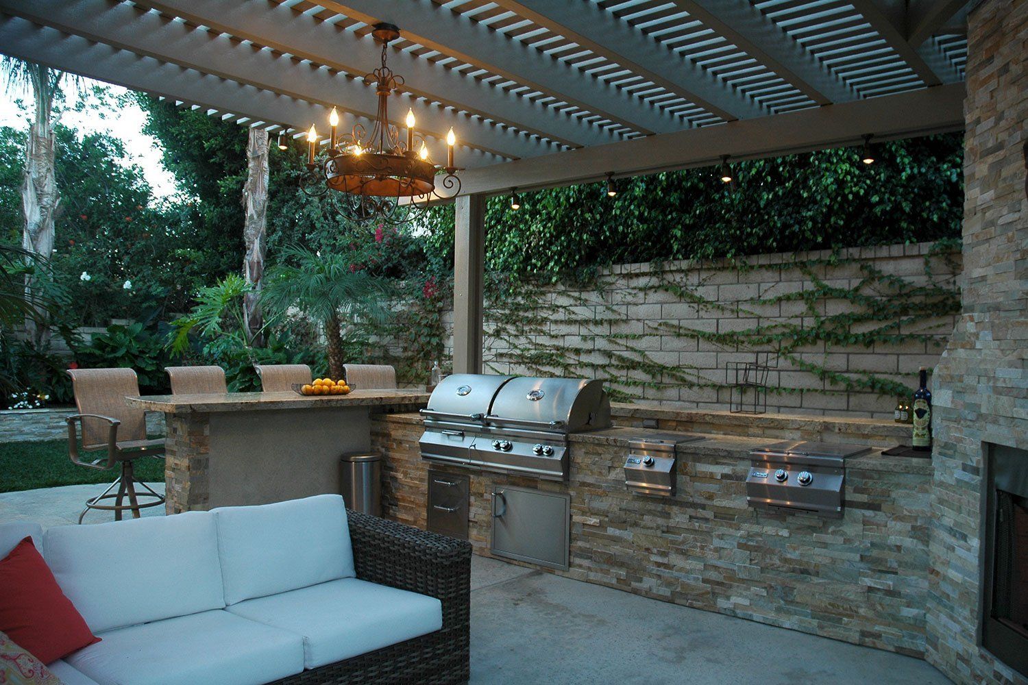 Covered patio with wicker seating, built-in grill, and dining table beside a stone wall and greenery