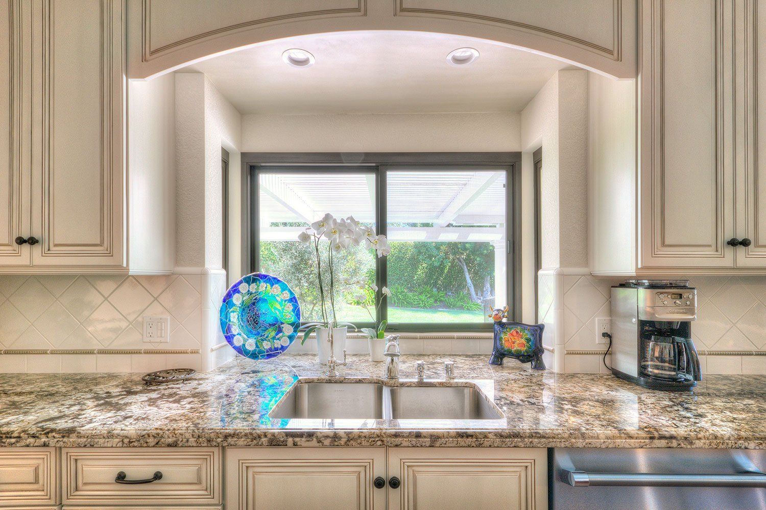 Kitchen sink with granite countertop, window, cabinets, and small blue decorative plate on the left.