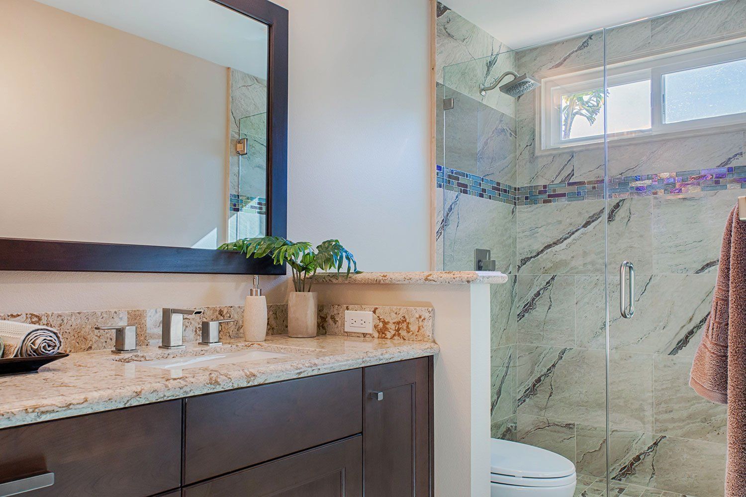 Modern bathroom vanity with mirror, granite countertop, and glass-enclosed shower with marble tile.