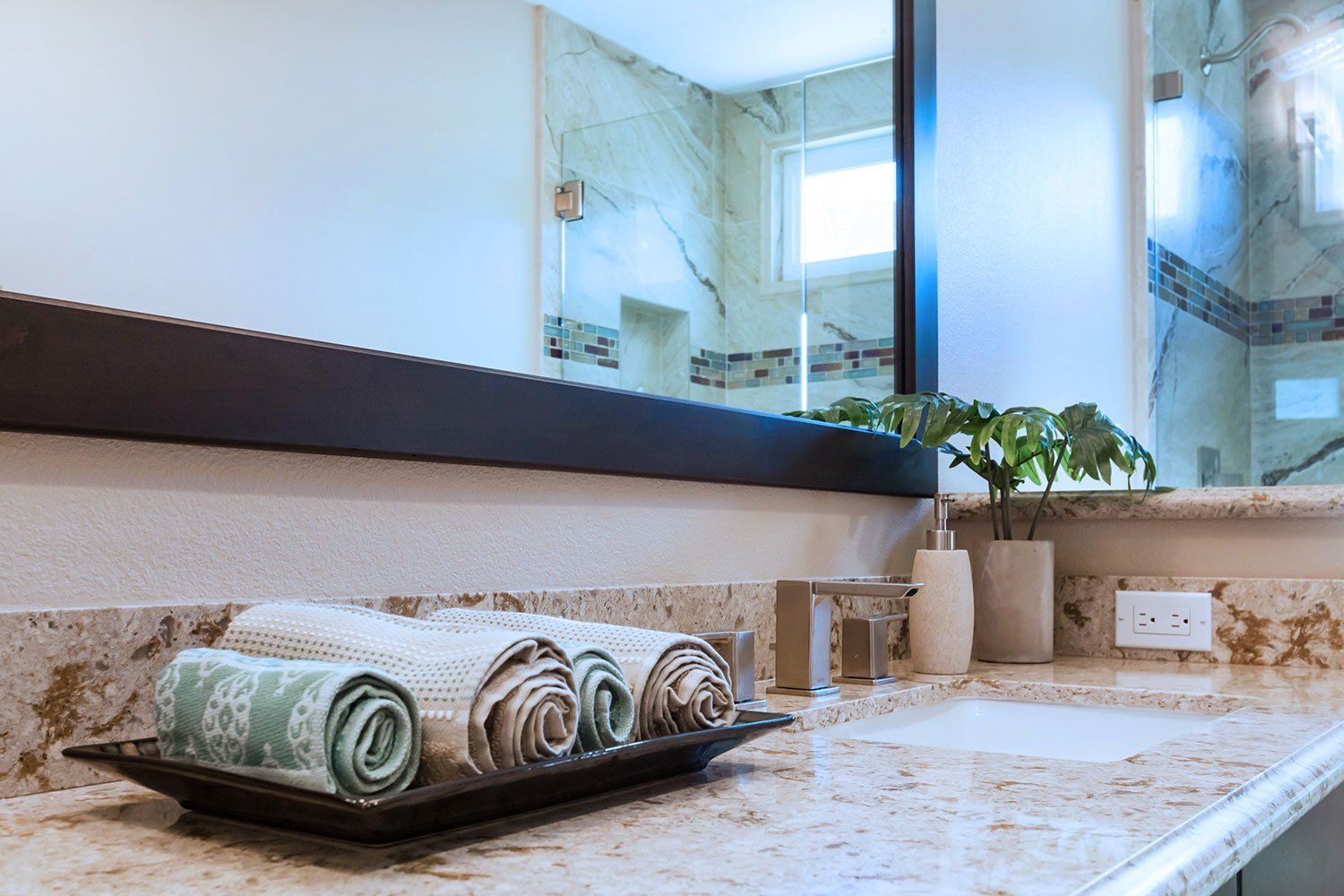 Bathroom countertop with rolled towels on a tray, beige stone surface, mirror, and sink accessories