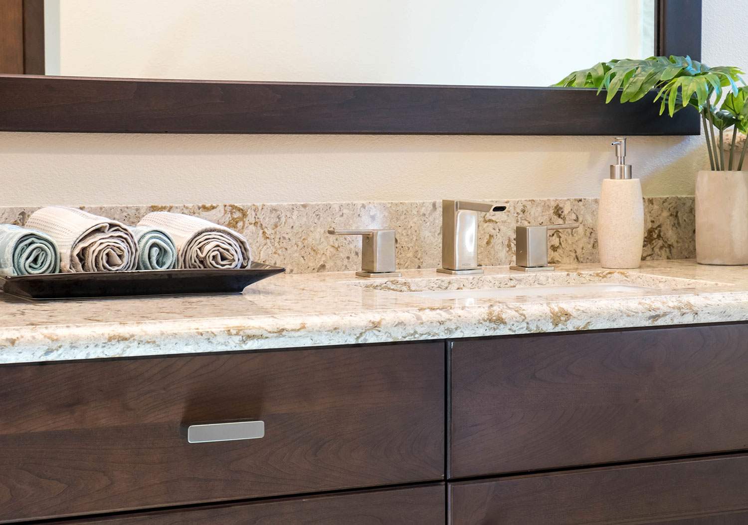 Modern bathroom vanity with dark wood cabinets, light stone countertop, tray of rolled towels, and plants
