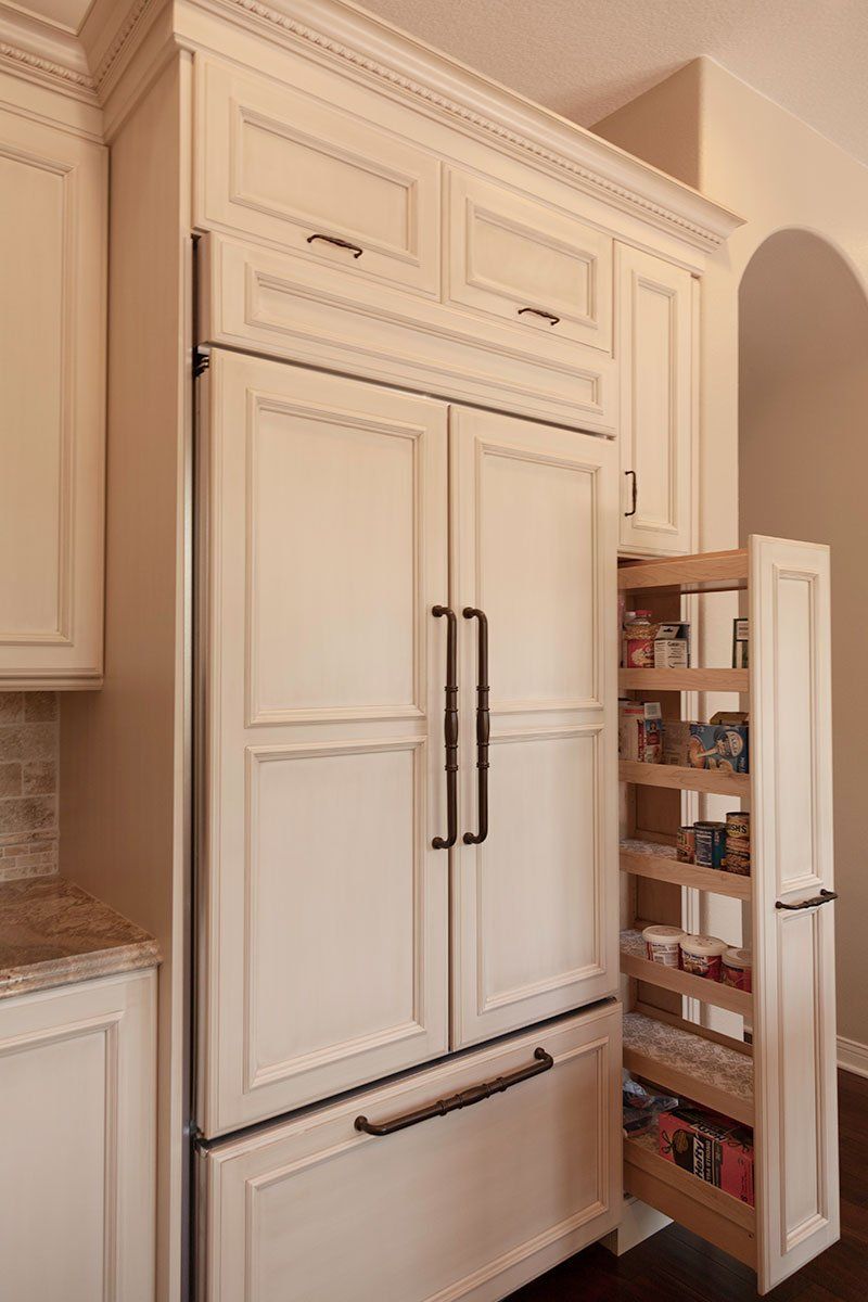 White built-in kitchen cabinetry with a double-door refrigerator and an open pantry door showing shelves of groceries.