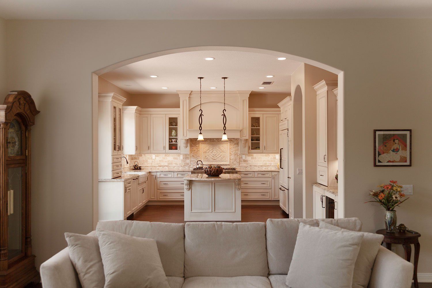 Bright living room opening to a white kitchen, with a beige sofa in the foreground and an arched doorway.