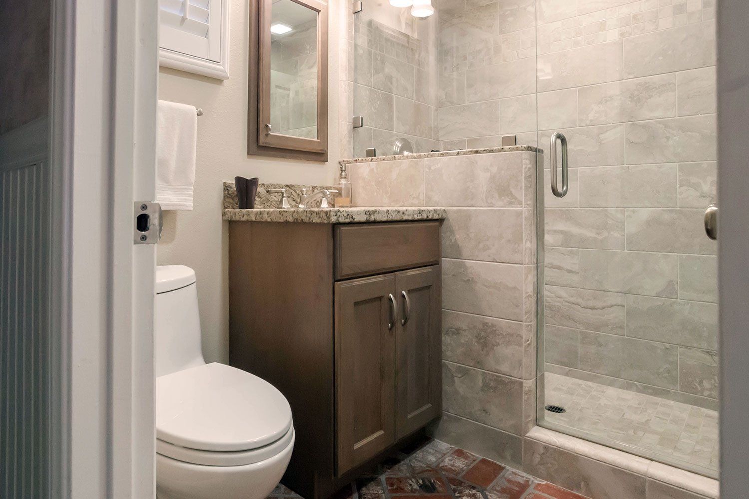Small bathroom with toilet, vanity, mirror, and glass shower, in beige and wood tones.