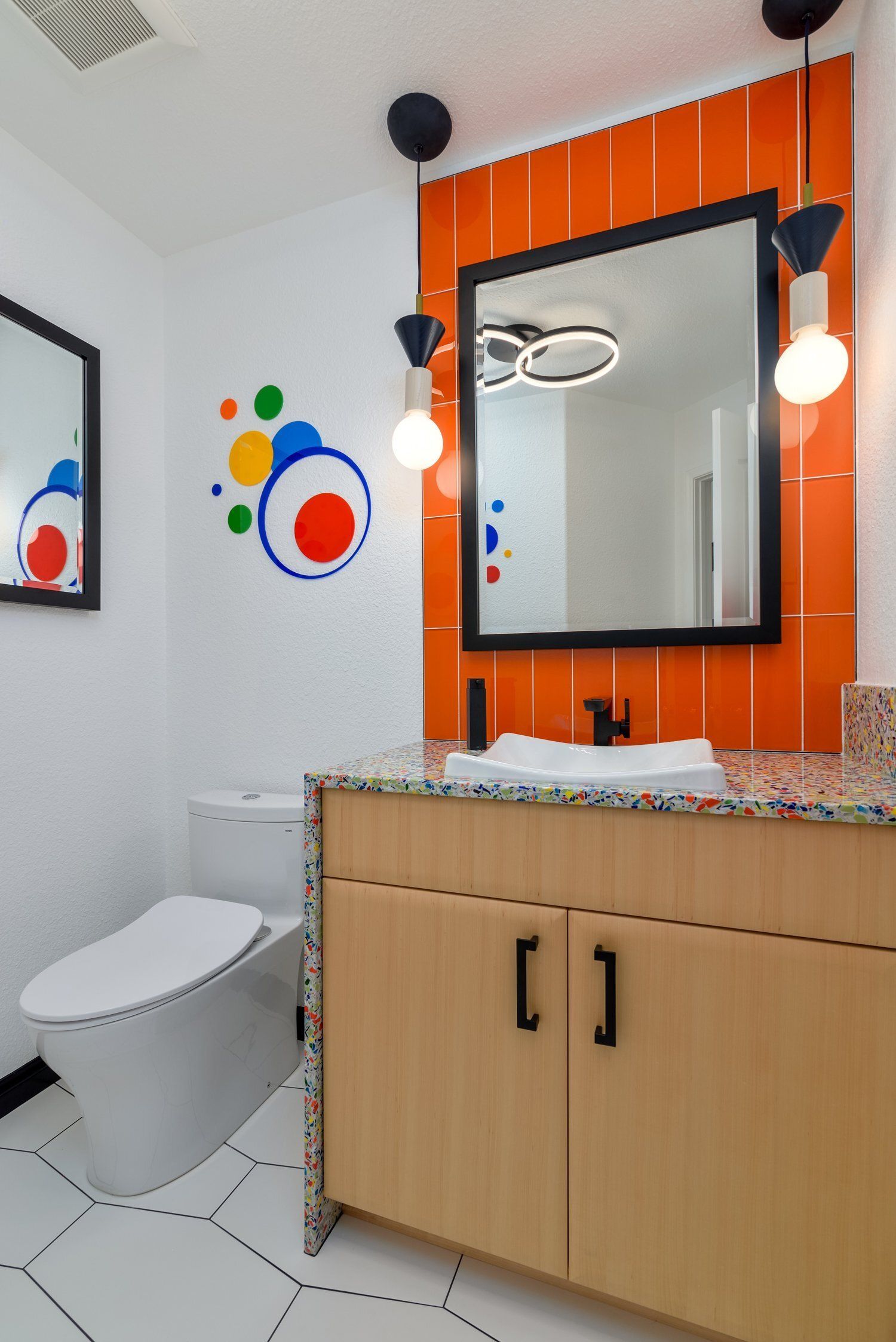 Modern bathroom with orange accent wall, round mirror, sink cabinet, toilet, and hanging lights