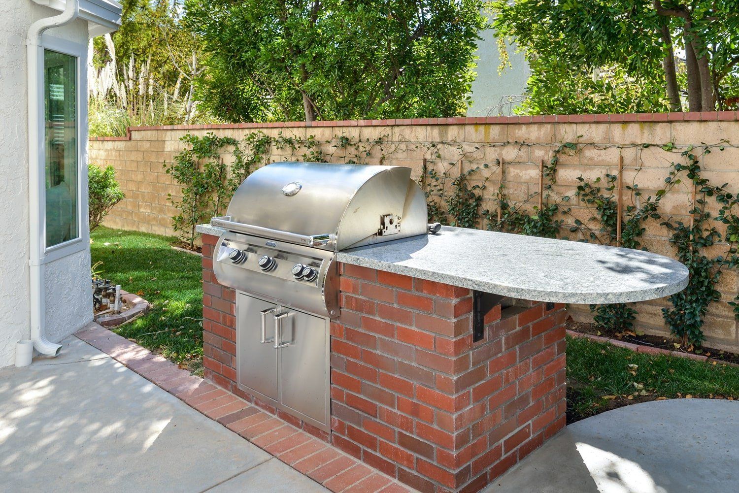 Outdoor built-in gas grill with brick base and stone side counter in a backyard patio.
