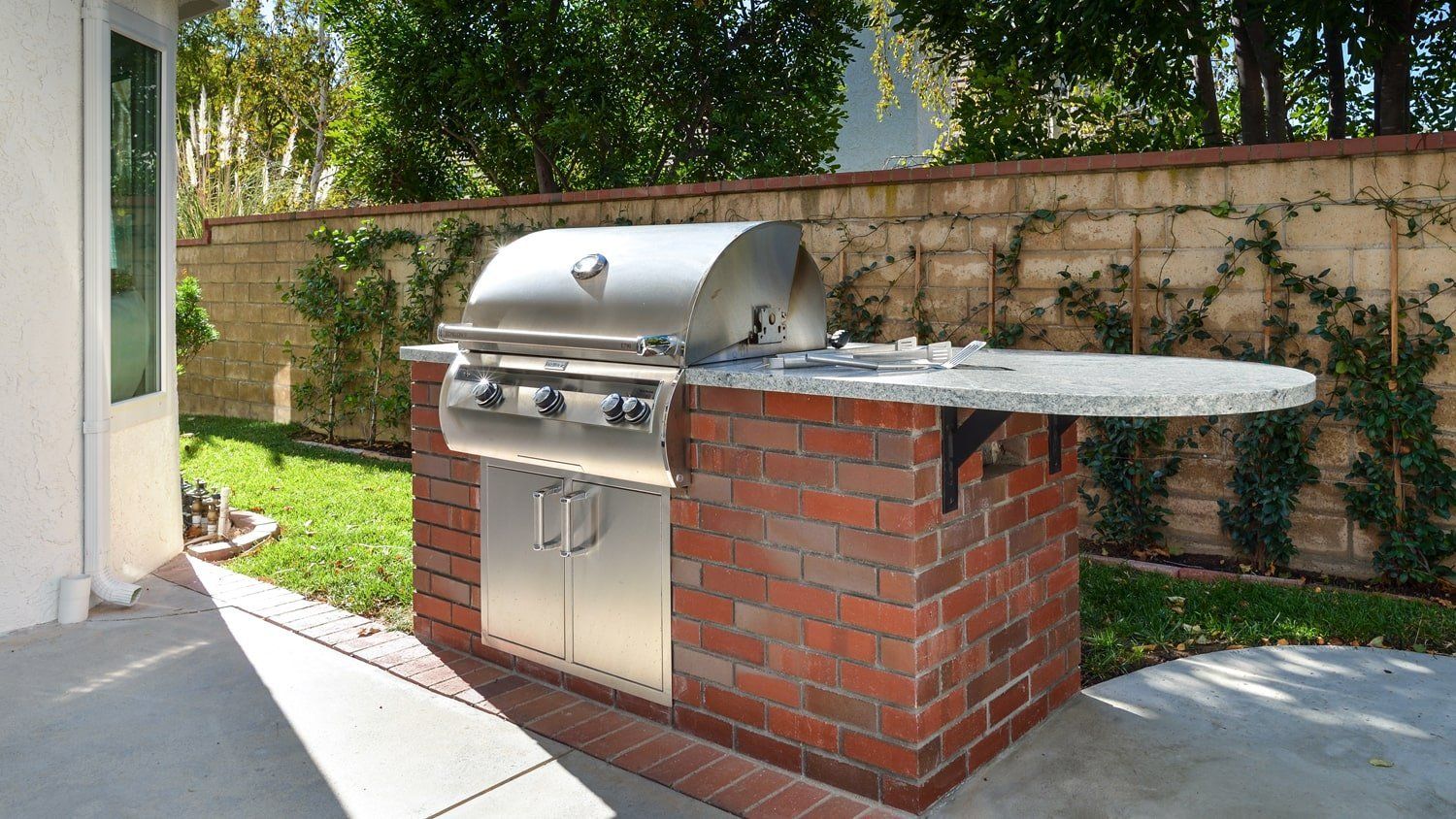 Outdoor stainless steel grill built into a red brick island beside a patio and stone wall.