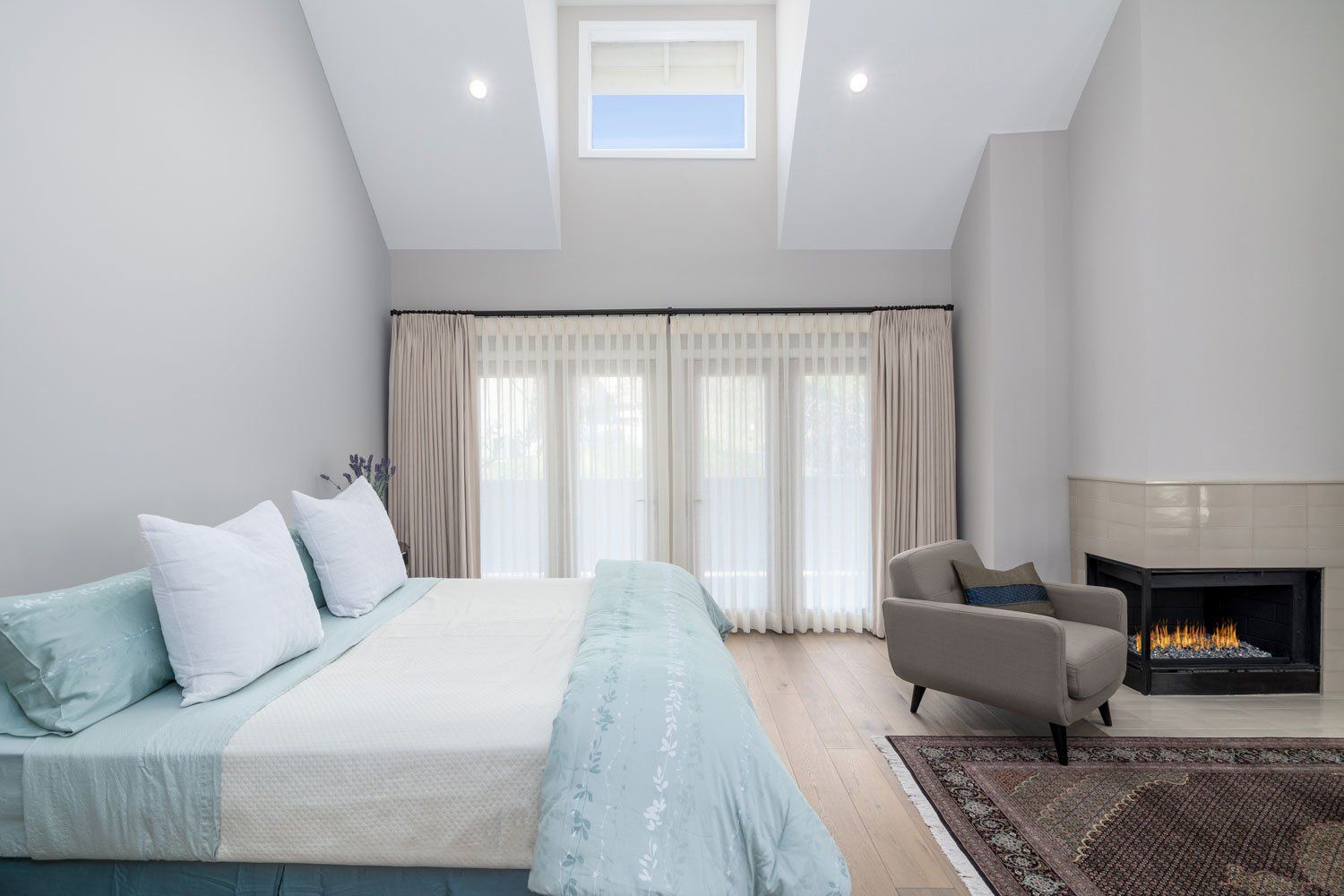Bright modern bedroom with a bed, armchair, fireplace, skylight, and sheer curtains.