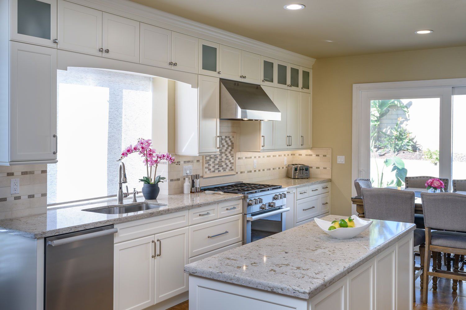 Bright modern kitchen with white cabinets, granite island, stainless range, and a small dining area.