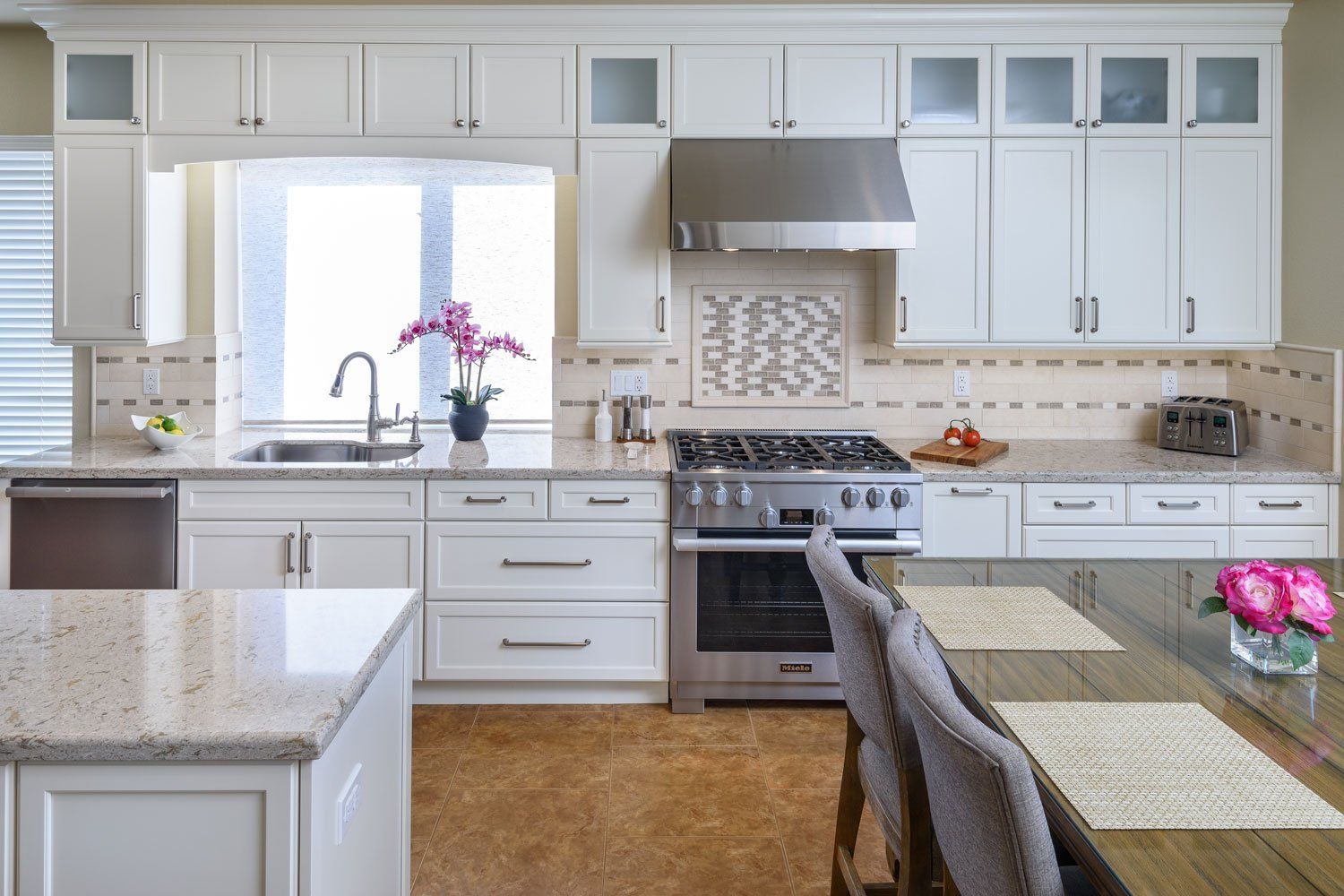 Bright white kitchen with island, granite countertops, stainless range hood, and pink flowers by the sink.