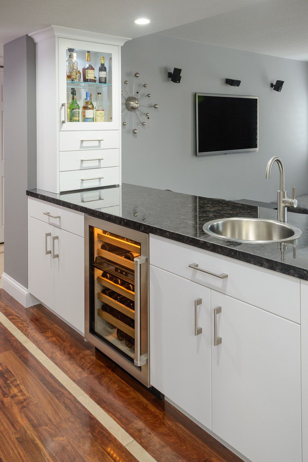 Modern kitchen bar with black granite countertop, sink, wine fridge, and built-in TV against gray wall