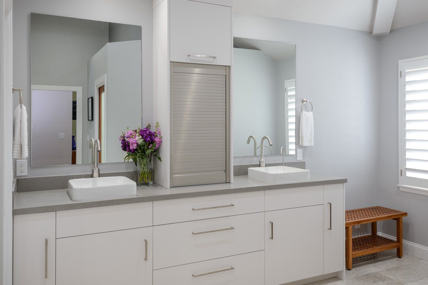 Modern white bathroom vanity with two mirrors, sinks, flowers, and window shutters