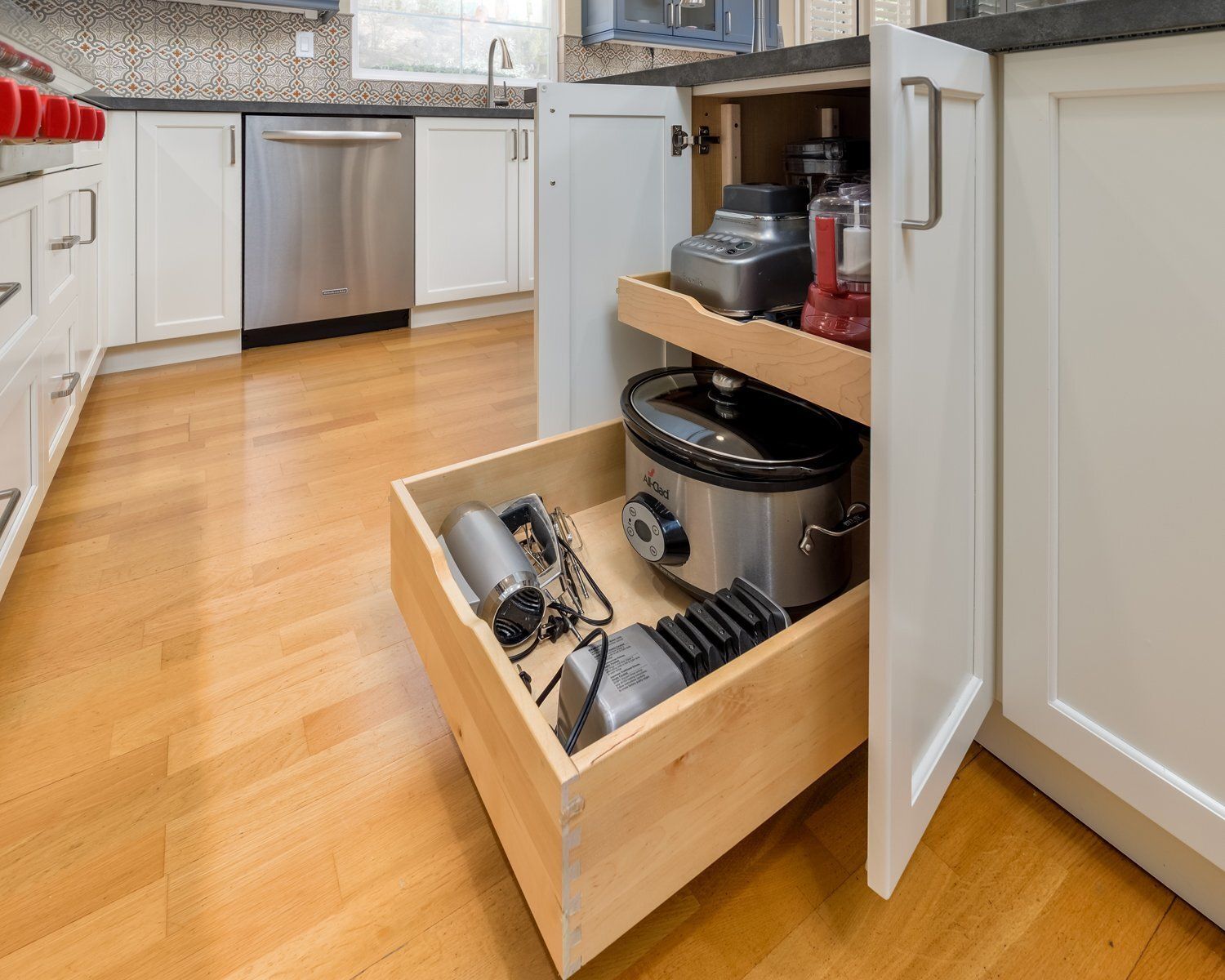 Open kitchen cabinet with pull-out drawers holding pots, pans, and appliances on a wooden floor.