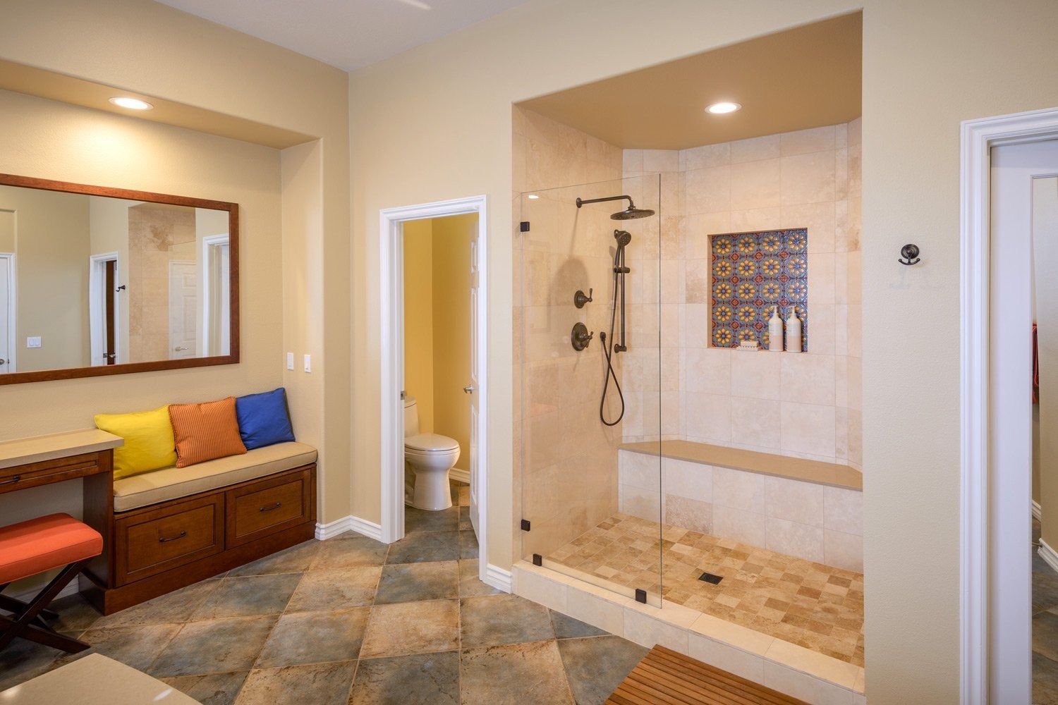 Accessible bathroom with walk-in shower, toilet, bench, and bright orange and blue cushions by a mirror.