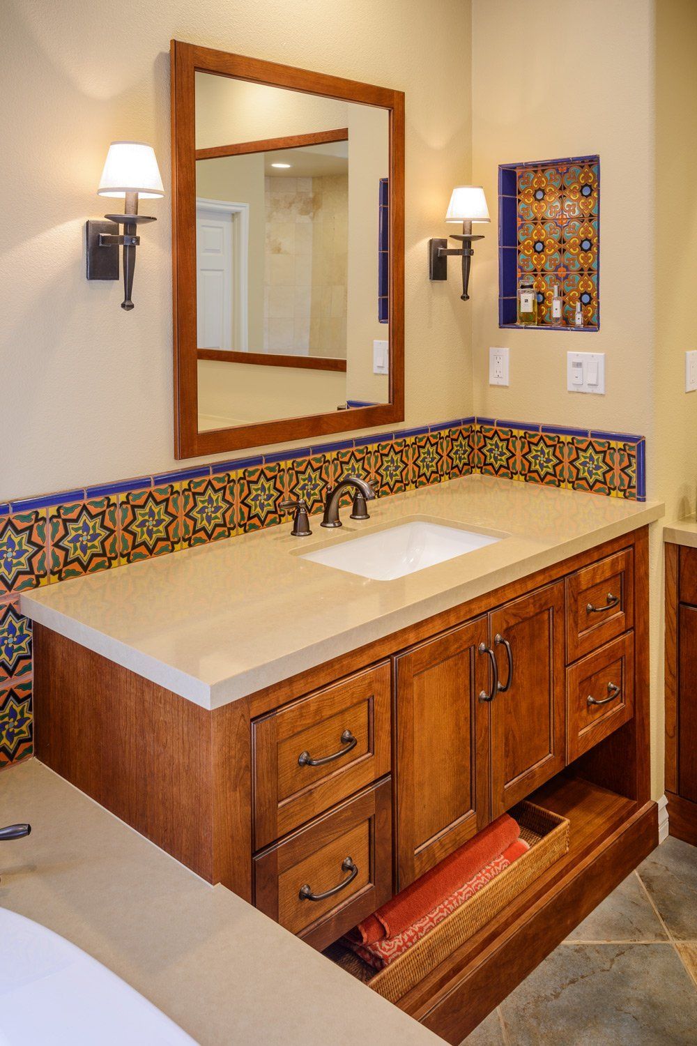 Bathroom vanity with wood cabinets, rectangular mirror, wall sconces, and tiled countertop with sink.