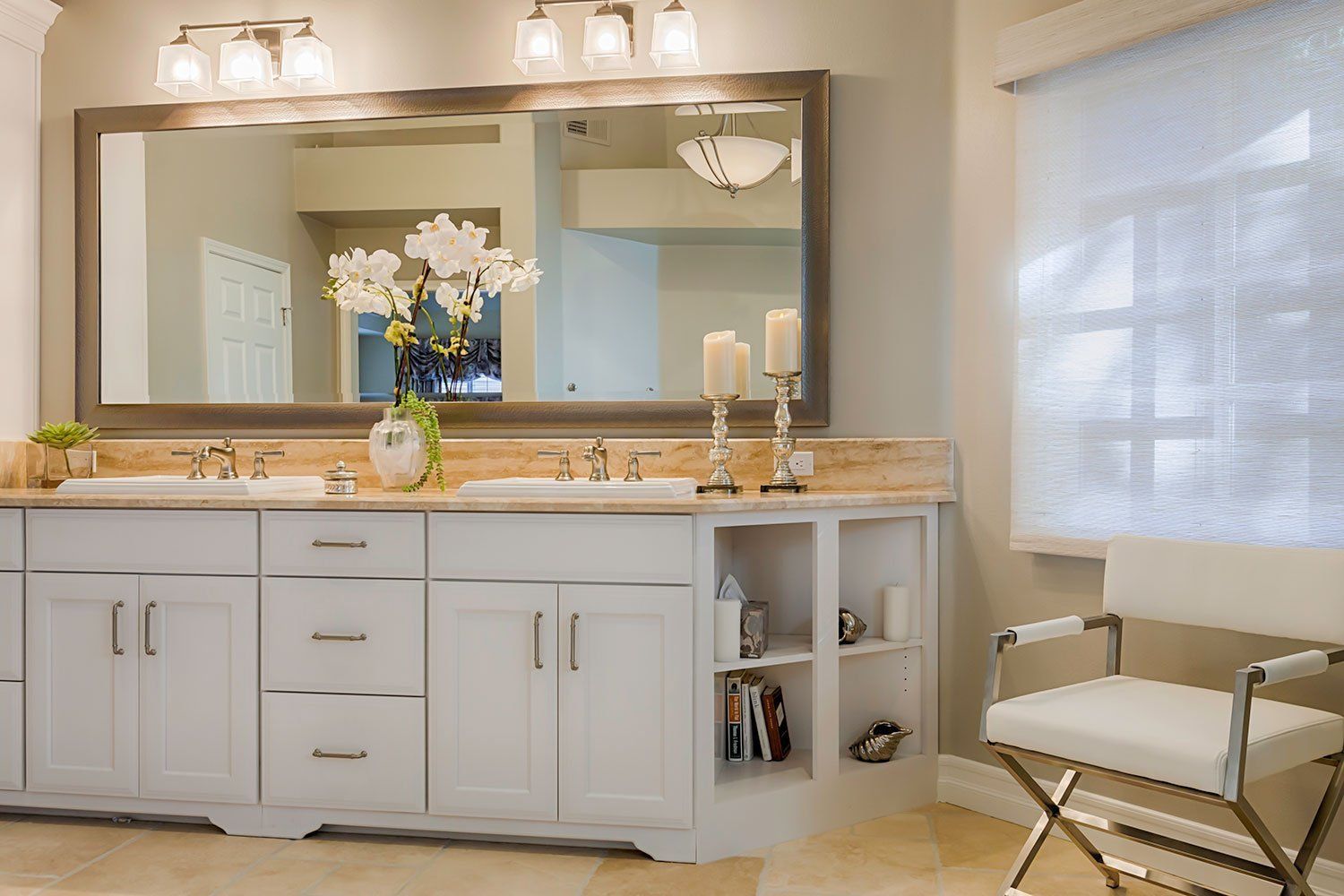 Bright bathroom vanity with double sinks, large mirror, flowers, and a folding chair by a window.