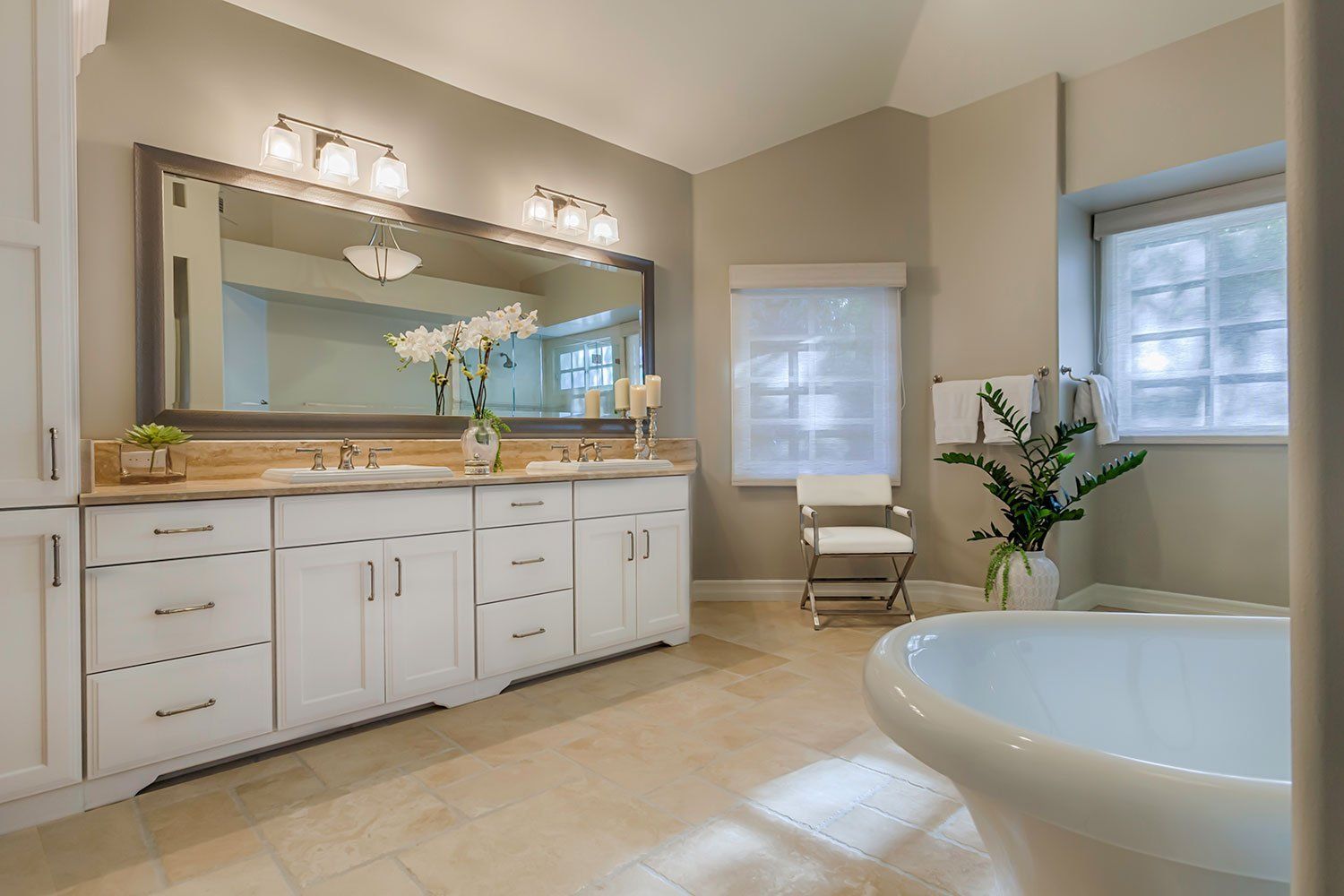 Bright modern bathroom with white vanity, large mirror, windows, chair, and a freestanding tub