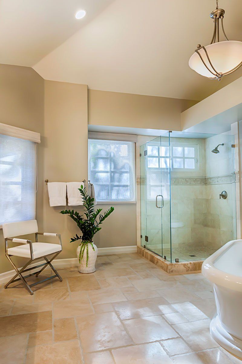 Bright beige bathroom with glass shower, tiled floor, windows, chair, plant, and chandelier lighting