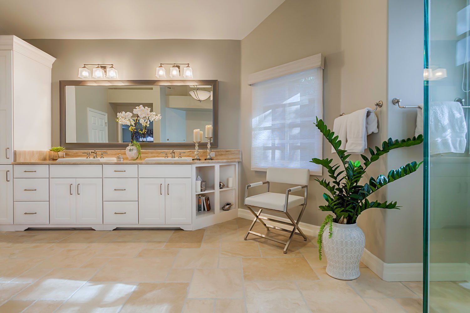 Bright bathroom with white vanity, large mirror, glass shower, and a potted plant near a folding chair.
