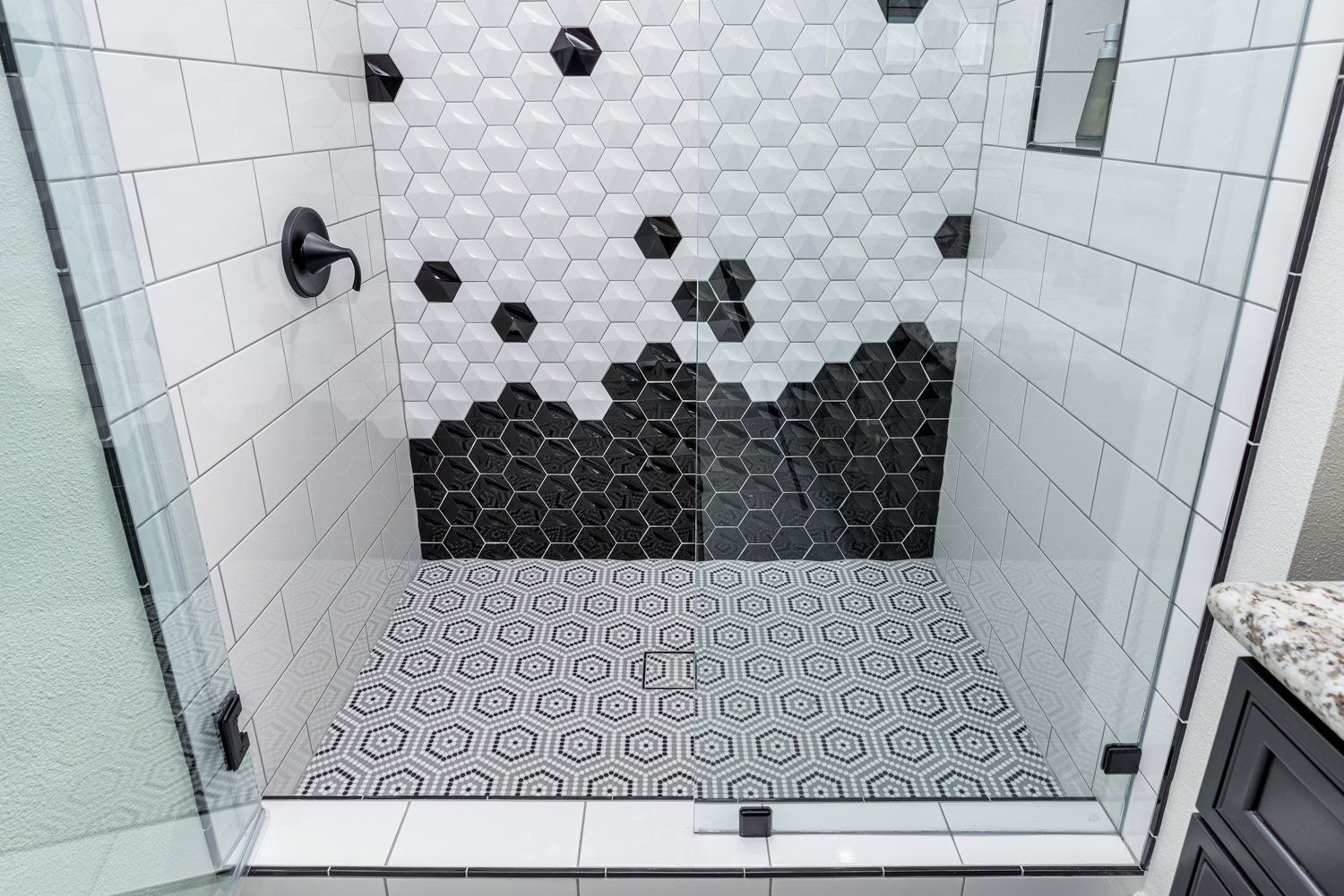 Modern shower stall with white hex tile, black accent wall, and mosaic floor behind glass door