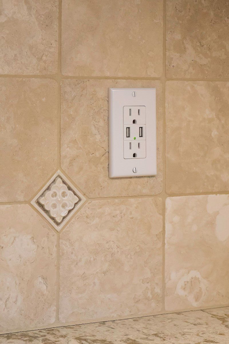 Beige tiled wall with a white electrical outlet and a small decorative tile accent.