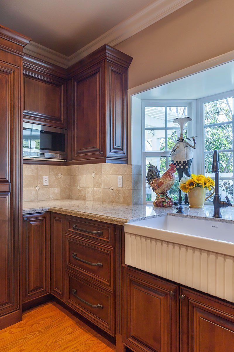 Wooden kitchen cabinets beside a sink under a sunlit window with flowers and a decorative vase