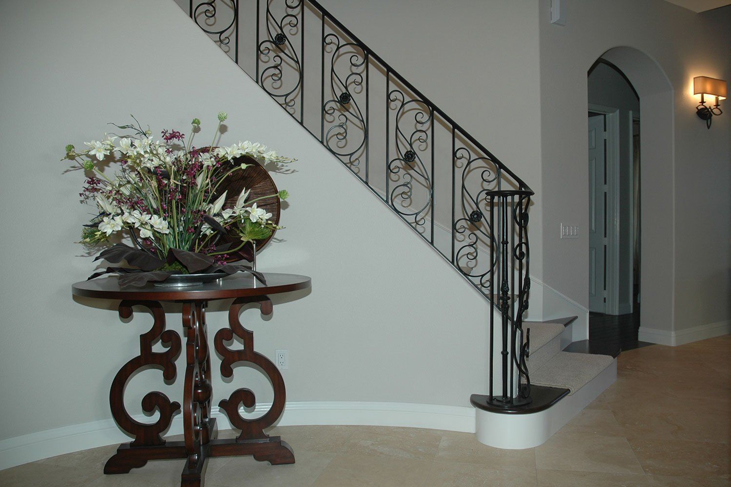 Staircase with ornate wrought-iron railing beside a decorative table and floral arrangement in a foyer.