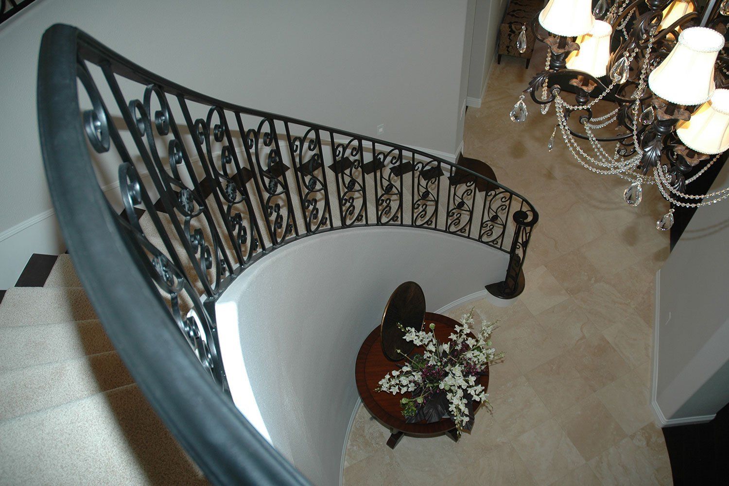 Curved staircase with ornate black railing, white wall, beige floor, and a floral arrangement below.