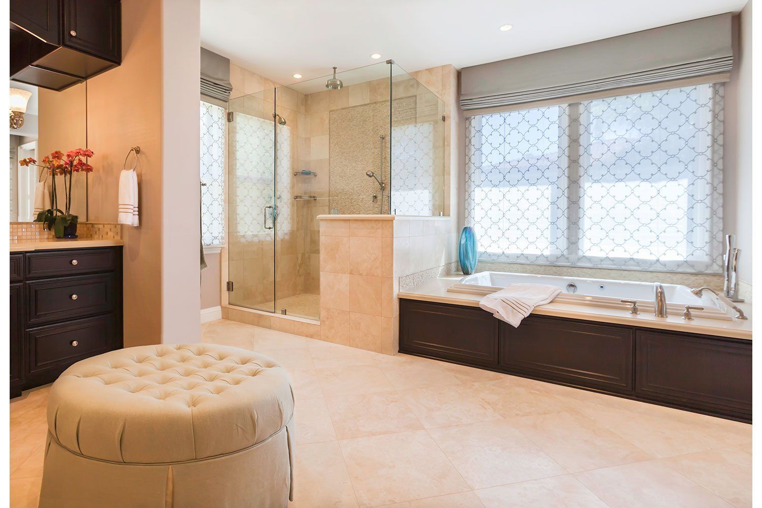 Modern bathroom with glass shower, soaking tub by window, dark vanity, and tufted ottoman