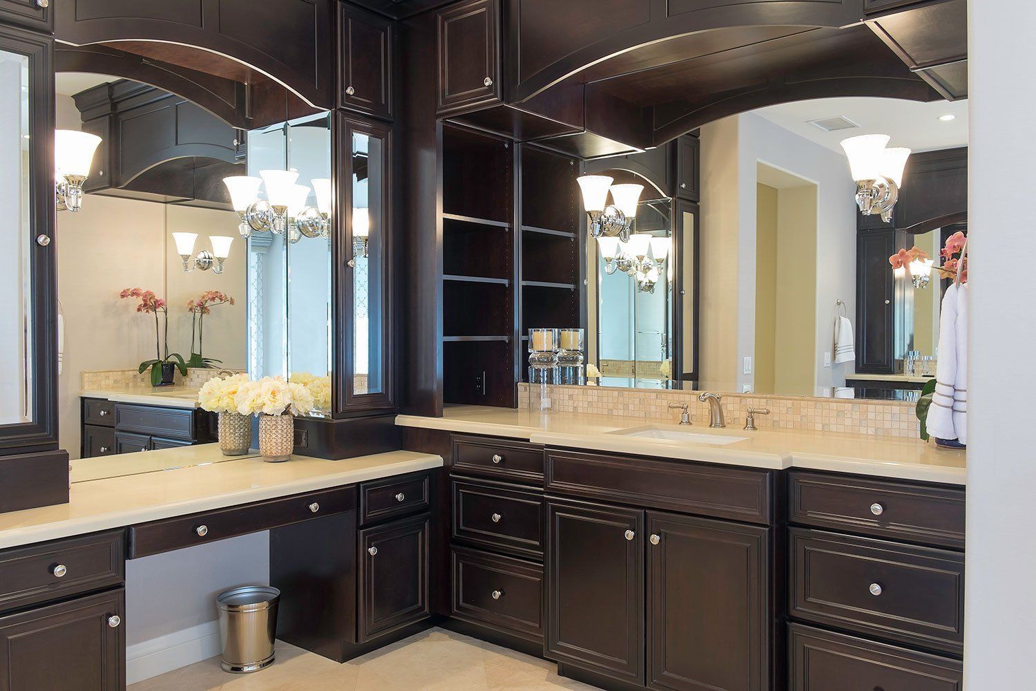 Elegant bathroom vanity with dark wood cabinets, large mirrors, beige countertop, and chandelier lighting.