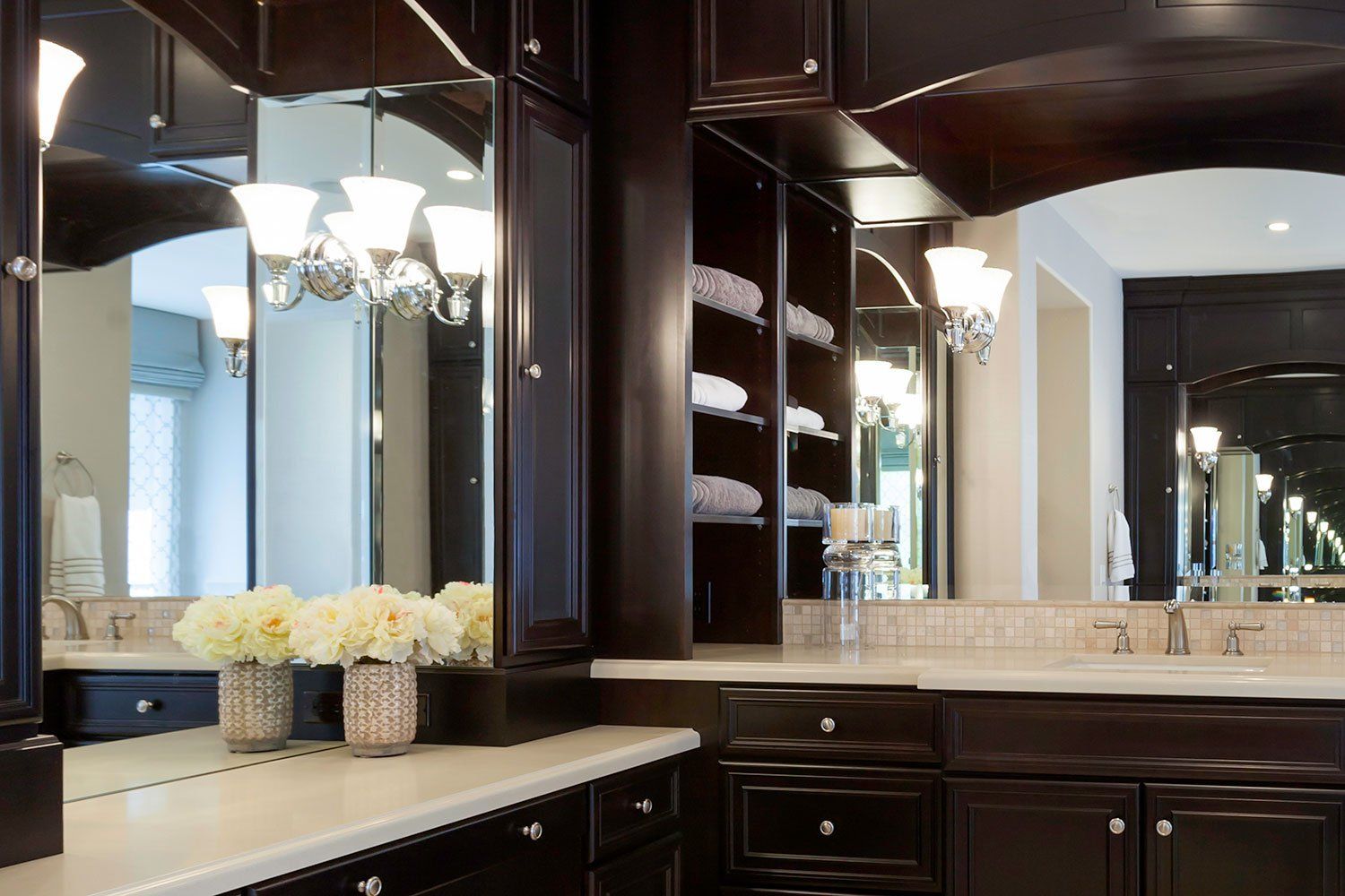 Dark wood bathroom vanity with double mirrors, cream countertop, and white floral arrangements