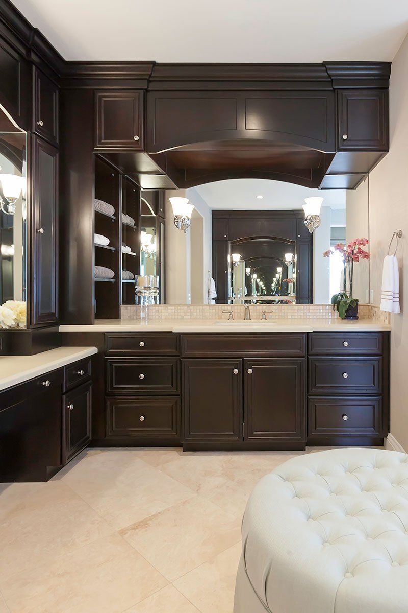 Elegant dark wood bathroom vanity with large mirror, beige tile floor, and white tufted ottoman.