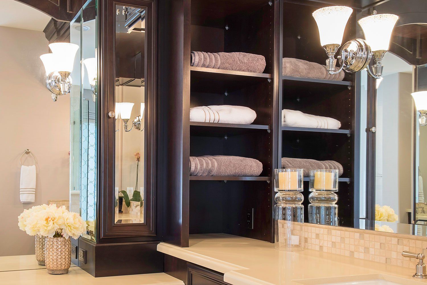 Bathroom vanity with dark wood cabinets, mirrors, lights, folded towels, and neatly arranged glass jars.